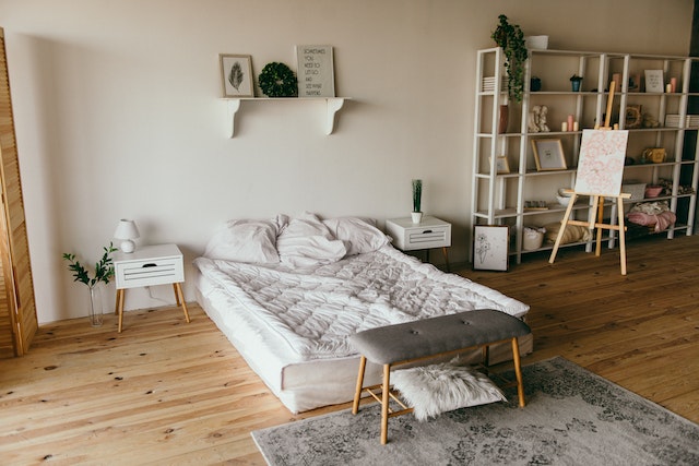 A minimalist bedroom in a rental property with a white bookshelf on the rights side of the bed which is on a blond hardwood floor and dressed with a white comforter